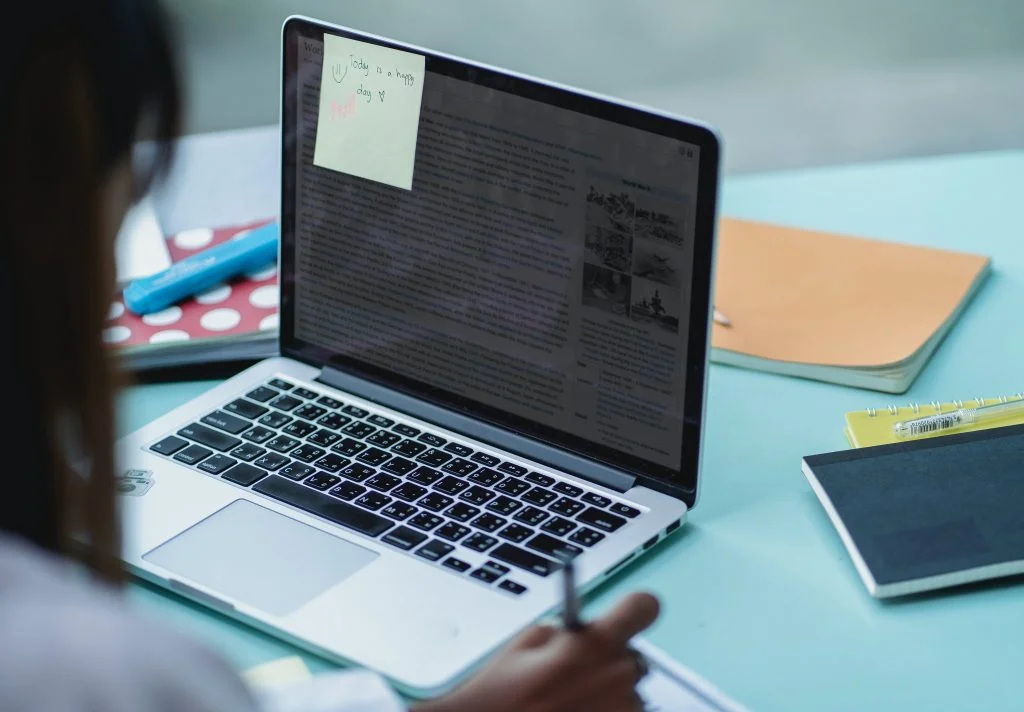 Eine Person nutzt einen Laptop mit einem Post-it-Zettel auf dem Bildschirm und schreibt in ein Notizbuch.