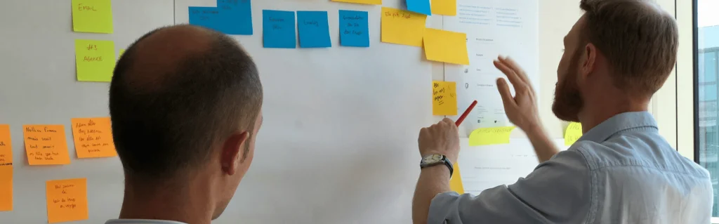 2 Männer stehen vor einem Whiteboard und setzen buntfarbige Post-its.